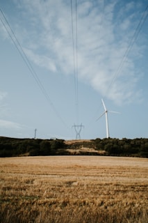 A wind turbine stands tall on a grassy hill under a partly cloudy sky. Power lines stretch across the scene, connecting to a distant pylon. The foreground features a golden field typical of harvested crops, surrounded by patches of green trees.
