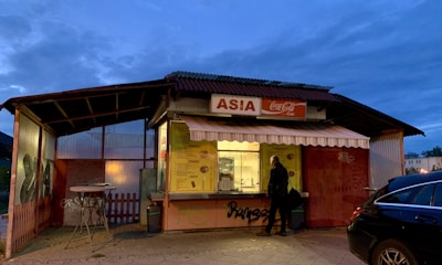 A small food kiosk with a sign labeled 'Asia' and a Coca-Cola logo, illuminated in the evening light. The kiosk has a menu displayed on the front and an awning above. A person stands in front of the counter, possibly ordering food. The surrounding area appears dimly lit with some graffiti on the side of the kiosk.