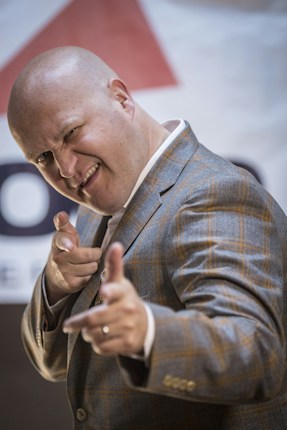 A bald man dressed in a plaid suit jacket is pointing both his fingers forward in a playful or confident manner, with a slight smile and a wink on his face. The background is blurred, possibly featuring some signage with partial text visible.