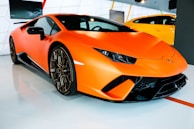 A sleek, orange sports car with a low, aerodynamic design is displayed in a showroom. The car has sharp lines, large black tires with bronze rims, and distinctive headlights. The background shows another vehicle in bright yellow and modern showroom decor with geometric patterns.