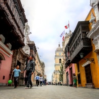 A bustling street scene in a city with colonial architecture, featuring people walking along a cobblestone path. The buildings are adorned with wooden balconies, and flags are seen atop one of the structures. The street is lined with colorful buildings, including ones painted in pink and yellow, contributing to the vibrant and historical ambiance.