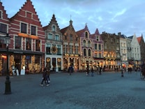 A view of a charming town square featuring a row of colorful, ornate buildings with steep gabled roofs and festive holiday decorations, such as lights and wreaths. The cobblestone street in front of the buildings is dotted with various people walking and socializing.