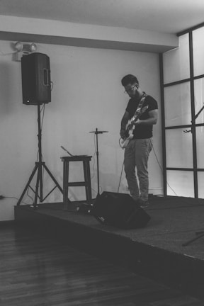 A person is standing on a small elevated platform playing an electric guitar. The room has minimal decor and is equipped with a tall speaker, a stool, and various musical equipment. The scene is captured in black and white.