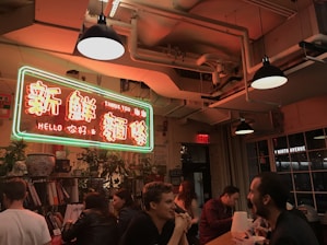 A warmly lit indoor setting featuring a neon sign with Chinese characters and English words reading 'Thank You' and 'Hello'. People are seated around a long table, engaging in conversation and eating. The ceiling has exposed pipes and industrial-style lighting, adding to the cozy ambiance. Potted plants and books are visible on shelves around the space.