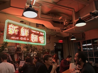 A warmly lit indoor setting featuring a neon sign with Chinese characters and English words reading 'Thank You' and 'Hello'. People are seated around a long table, engaging in conversation and eating. The ceiling has exposed pipes and industrial-style lighting, adding to the cozy ambiance. Potted plants and books are visible on shelves around the space.