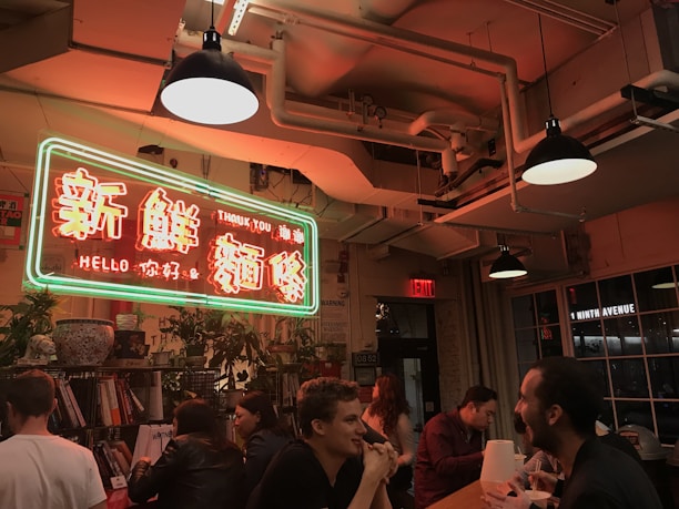 A warmly lit indoor setting featuring a neon sign with Chinese characters and English words reading 'Thank You' and 'Hello'. People are seated around a long table, engaging in conversation and eating. The ceiling has exposed pipes and industrial-style lighting, adding to the cozy ambiance. Potted plants and books are visible on shelves around the space.