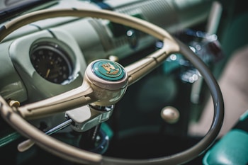 A vintage car steering wheel is centrally featured, showcasing its classic design with a distinctive emblem in the center. The dashboard exhibits an analog speedometer and smooth, retro-style details.
