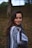 A young woman stands outdoors, wrapped in a patterned poncho. The background features a blurred grassy area and trees, indicating a natural setting during autumn. The woman has long, dark hair and a soft smile.