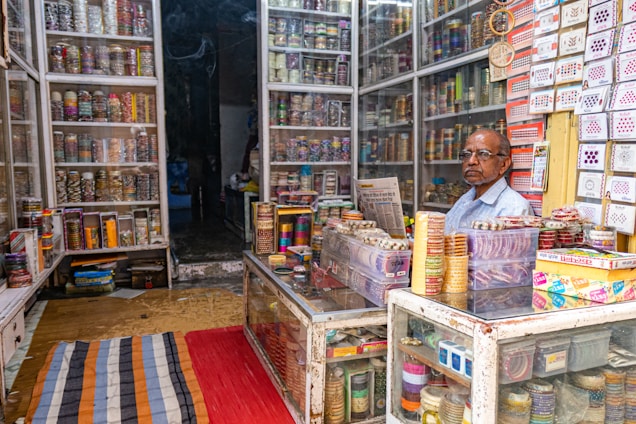 Inside a colorful shop filled with glass bangles neatly displayed on shelves. A man sits behind a counter reading a newspaper, surrounded by stacks of bangles and other small items. Brightly colored rugs are placed on the floor.