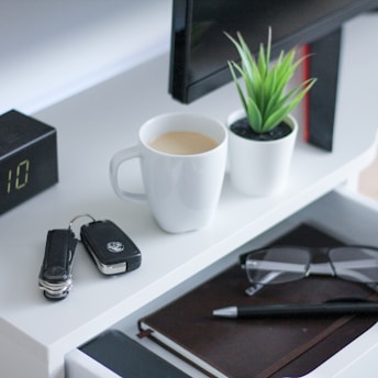 On a white desk, there is a mug filled with coffee, a small potted plant with green leaves, a pair of eyeglasses, a black pen, a closed notebook, and a set of car keys with a remote fob. Nearby, a digital clock displays the time as 1:10.