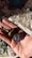 A hand holding two polished stones, one with a shiny brown stripe pattern resembling tiger's eye and the other a smooth purple stone possibly amethyst, set against a background scattered with various unpolished crystals and stones.