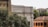 An architectural scene featuring the Accademia di Belle Arti building with classic columns and ornate masonry. Surrounding the building, lush greenery and trees provide a natural foreground, while nearby structures with tiled roofs add to the historical atmosphere.