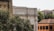 An architectural scene featuring the Accademia di Belle Arti building with classic columns and ornate masonry. Surrounding the building, lush greenery and trees provide a natural foreground, while nearby structures with tiled roofs add to the historical atmosphere.