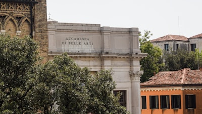 An architectural scene featuring the Accademia di Belle Arti building with classic columns and ornate masonry. Surrounding the building, lush greenery and trees provide a natural foreground, while nearby structures with tiled roofs add to the historical atmosphere.