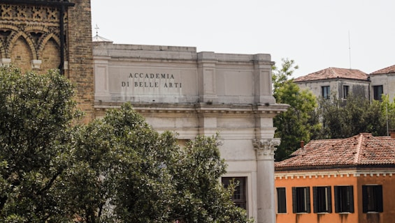 An architectural scene featuring the Accademia di Belle Arti building with classic columns and ornate masonry. Surrounding the building, lush greenery and trees provide a natural foreground, while nearby structures with tiled roofs add to the historical atmosphere.