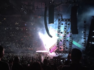 A large, energetic concert audience fills an indoor arena, with stage lights and smoke creating a dynamic atmosphere. The stage is equipped with towering scaffolding and colorful lights. Many people in the crowd are capturing the moment with their phones.