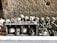 A collection of human skulls and bones is arranged in an orderly fashion against a rough, stone backdrop. Several skulls are placed inside a box, adorned with rosaries, flowers, and other small offerings or memorabilia. A bouquet of artificial roses is visible alongside the skulls, adding a poignant touch.