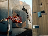 A modern architectural scene featuring abstract and angular building facades. The structures are composed of geometric shapes and metallic surfaces, reflecting light in varied directions. A cherry picker with a worker in a red safety harness is elevated near the building. There is a pole with a banner reading 'IDEAS' attached to it.