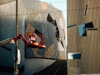 A modern architectural scene featuring abstract and angular building facades. The structures are composed of geometric shapes and metallic surfaces, reflecting light in varied directions. A cherry picker with a worker in a red safety harness is elevated near the building. There is a pole with a banner reading 'IDEAS' attached to it.