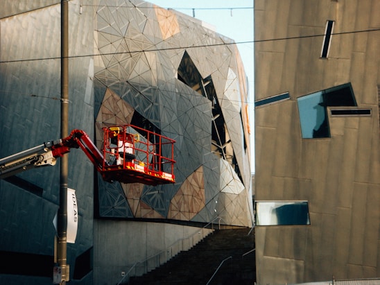 A modern architectural scene featuring abstract and angular building facades. The structures are composed of geometric shapes and metallic surfaces, reflecting light in varied directions. A cherry picker with a worker in a red safety harness is elevated near the building. There is a pole with a banner reading 'IDEAS' attached to it.