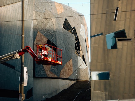 A modern architectural scene featuring abstract and angular building facades. The structures are composed of geometric shapes and metallic surfaces, reflecting light in varied directions. A cherry picker with a worker in a red safety harness is elevated near the building. There is a pole with a banner reading 'IDEAS' attached to it.