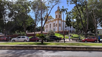 A historic church is situated in a lush, green park setting. The building features classic architectural style with a white facade and orange border details. Trees surround the area, casting shade over the parked cars and people walking by. The sky is clear and blue, adding a serene backdrop to the scene.