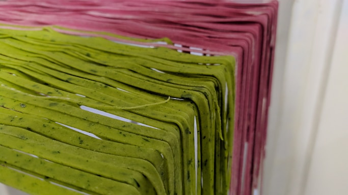 Racks of pasta are hanging side by side, with varying colors indicating different types or flavors. The pasta is neatly arranged and appears to be freshly made, showcasing green and pink hues.