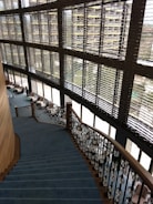 A modern interior featuring a large staircase with blue carpeting and ornate railings. The stairs descend alongside expansive windows with horizontal blinds, allowing natural light to fill the space. Comfortable chairs and small tables are located on the floor adjacent to the windows, fostering a relaxed seating area.