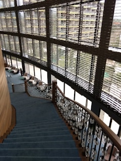 A modern interior featuring a large staircase with blue carpeting and ornate railings. The stairs descend alongside expansive windows with horizontal blinds, allowing natural light to fill the space. Comfortable chairs and small tables are located on the floor adjacent to the windows, fostering a relaxed seating area.