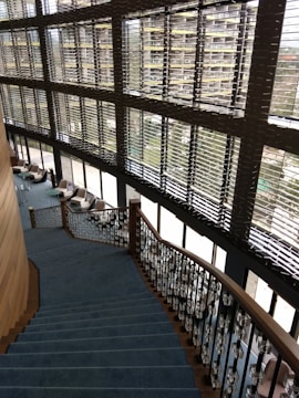 A modern interior featuring a large staircase with blue carpeting and ornate railings. The stairs descend alongside expansive windows with horizontal blinds, allowing natural light to fill the space. Comfortable chairs and small tables are located on the floor adjacent to the windows, fostering a relaxed seating area.