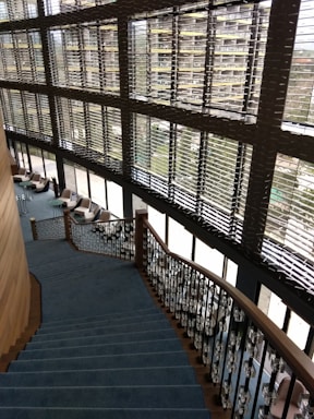 A modern interior featuring a large staircase with blue carpeting and ornate railings. The stairs descend alongside expansive windows with horizontal blinds, allowing natural light to fill the space. Comfortable chairs and small tables are located on the floor adjacent to the windows, fostering a relaxed seating area.
