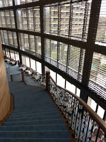 A modern interior featuring a large staircase with blue carpeting and ornate railings. The stairs descend alongside expansive windows with horizontal blinds, allowing natural light to fill the space. Comfortable chairs and small tables are located on the floor adjacent to the windows, fostering a relaxed seating area.