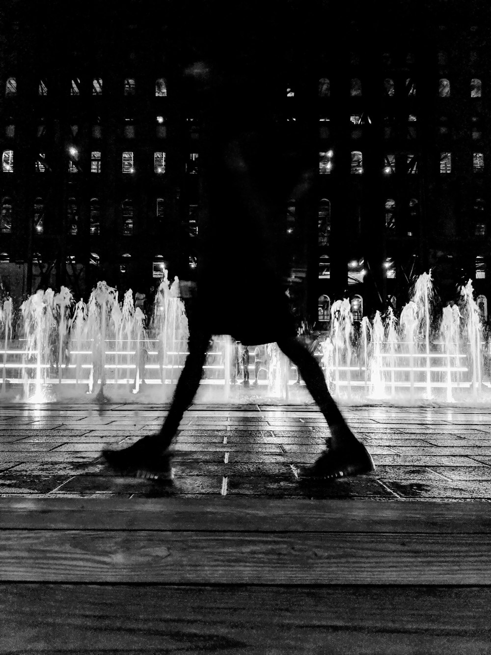 a black and white photo of a person walking in front of a fountain