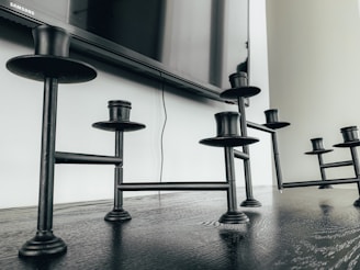 Black metal candle holders are placed on a dark wooden surface. Above them, a flat-screen television is affixed to a white wall. A power cable is visible extending down from the television.