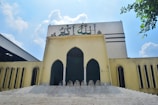 A large, beige-colored building with Arabic calligraphy on top. The building features three arched doorways and symmetrical rectangular windows flanking the sides. A set of wide stairs leads up to the entrance. The sky is clear with scattered clouds, and there are green leaves visible in the corner.