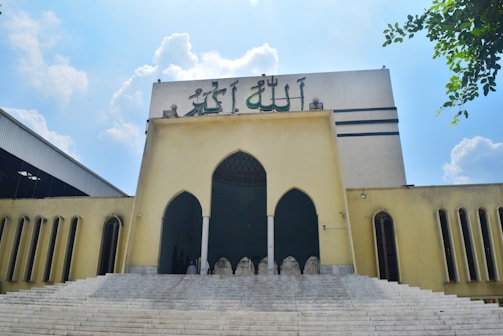A large, beige-colored building with Arabic calligraphy on top. The building features three arched doorways and symmetrical rectangular windows flanking the sides. A set of wide stairs leads up to the entrance. The sky is clear with scattered clouds, and there are green leaves visible in the corner.