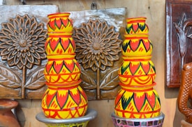 Two stacks of colorful clay pots are placed against decorative floral wall hangings. The pots are vibrantly painted with bold patterns in red, yellow, and black. The wall decor features raised flower designs with leaves, all in earthy brown tones.