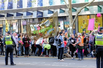 A large group of protesters are gathered on an urban street, holding signs and banners. The focus is on a banner with the words 'Disco Disruption' written on it. Police officers in uniform stand nearby, monitoring the situation. The crowd includes people of diverse ages, some holding flags and signs with symbols related to activism. There is a storefront in the background with large glass windows, displaying sports advertisements.