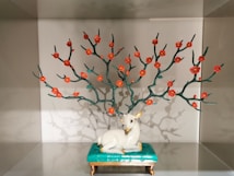 A decorative sculpture features a white deer sitting on a green pedestal. Above the deer, there are artistic branches with red flowers, creating a visually striking arrangement. The sculpture is showcased in a display box with soft lighting.