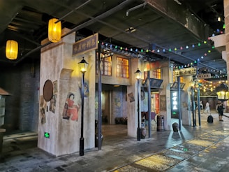 A street scene in a retro-styled indoor setting featuring vintage shop fronts with signs in Chinese characters, decorative windows, and traditional style street lamps. Posters of women in traditional attire are visible on the walls, and the area is adorned with colorful string lights. Cobbled flooring and historic architecture elements suggest an old-town atmosphere.