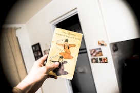A hand holds a vintage-style book titled 'Poems of the Late Tang'. The cover features an illustration of two historical figures in traditional Chinese attire. The background shows part of a room with a curtain, a doorway, and some pictures on a wall.