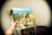A hand holds a vintage vinyl record cover with a blue and green design featuring 'The Sound of Music'. The cover displays an image of a group of children and a woman sitting on a staircase, reminiscent of a scene from the musical. In the background, a fedora is hanging on the wall, and there is furniture such as a chair and a shelf with stacked items.