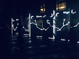 Illuminated constellation signs and symbols appear on glass panels, each featuring zodiac names and date ranges. The panels glow with bright outlines depicting stars and connecting lines against a dark backdrop. Surrounding reflective surfaces suggest a nighttime urban setting.