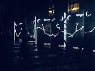 Illuminated constellation signs and symbols appear on glass panels, each featuring zodiac names and date ranges. The panels glow with bright outlines depicting stars and connecting lines against a dark backdrop. Surrounding reflective surfaces suggest a nighttime urban setting.