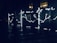 Illuminated constellation signs and symbols appear on glass panels, each featuring zodiac names and date ranges. The panels glow with bright outlines depicting stars and connecting lines against a dark backdrop. Surrounding reflective surfaces suggest a nighttime urban setting.