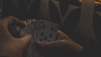 Two hands are holding a fan of cards with numbers and tree symbols against a dimly lit background. The shadows create a mysterious atmosphere, partially obscuring some details.