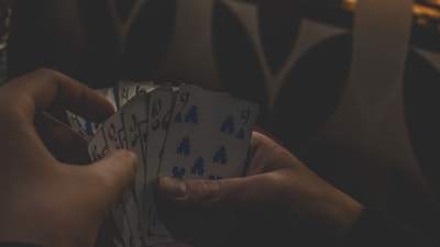 Two hands are holding a fan of cards with numbers and tree symbols against a dimly lit background. The shadows create a mysterious atmosphere, partially obscuring some details.