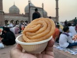 A hand holds a small white dish containing a spiral-shaped fried sweet. In the background, a large historic mosque with domes and minarets is visible, and many people are gathered around chatting and sitting on the ground.