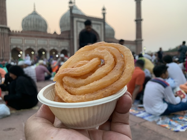 A hand holds a small white dish containing a spiral-shaped fried sweet. In the background, a large historic mosque with domes and minarets is visible, and many people are gathered around chatting and sitting on the ground.
