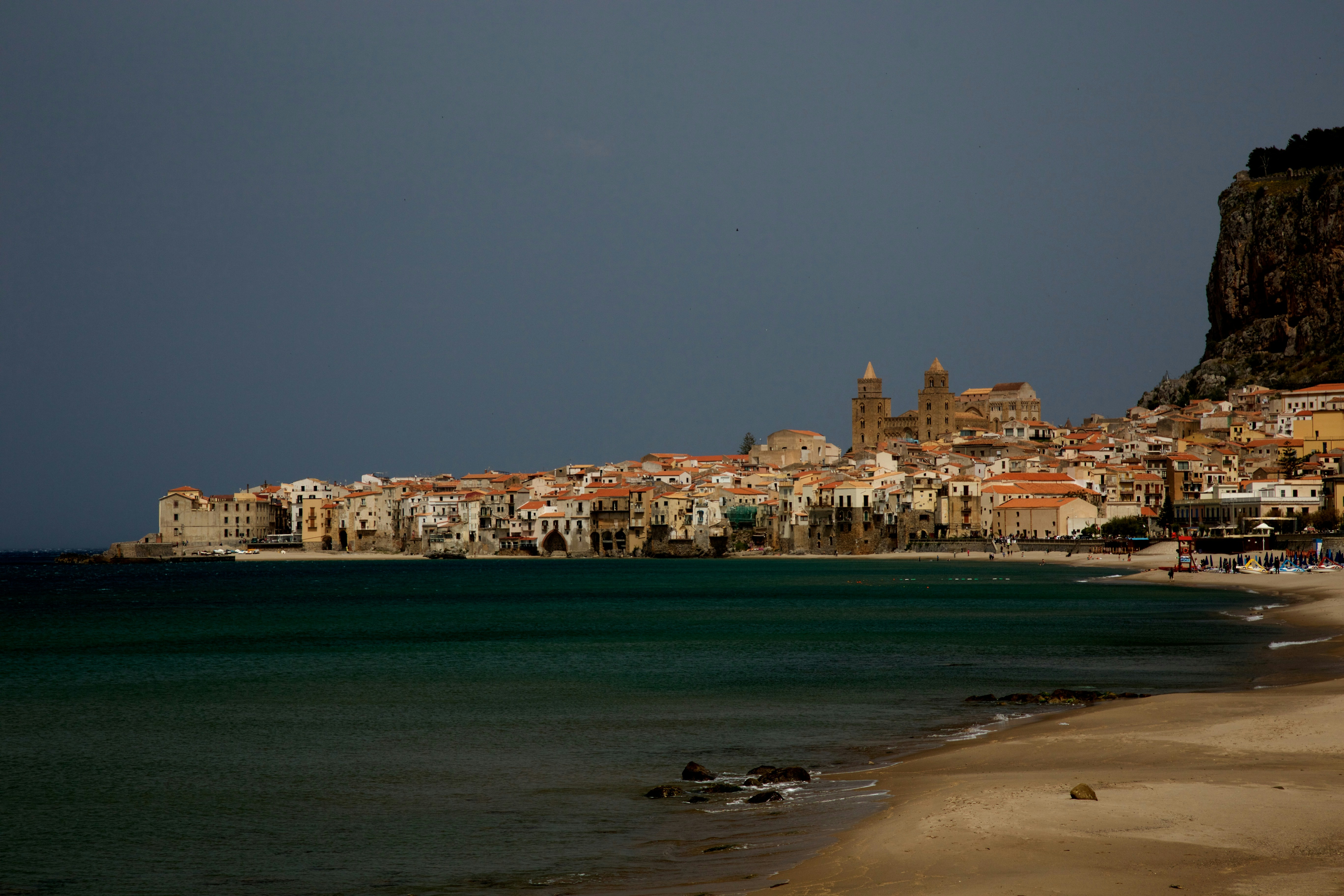 Mallorca coastal village at golden hour with terracotta rooftops, dark rocky cliffs, moody Mediterranean light